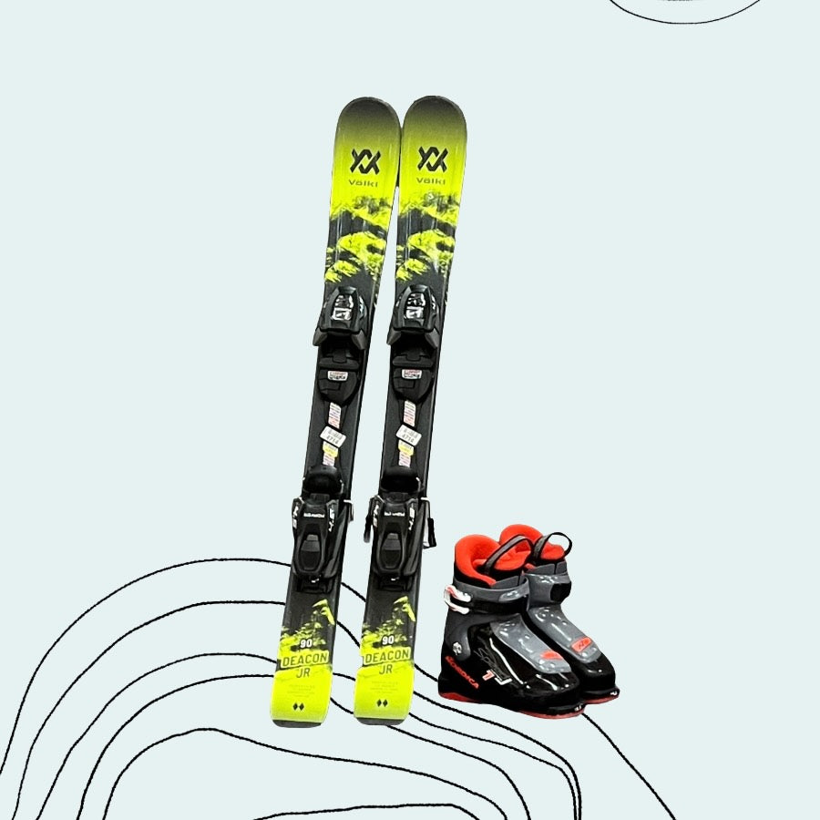 Pair of Toddler -sized yellow & black Volkl skis with black bindings and a black & red Nordica ski boot on a light blue background